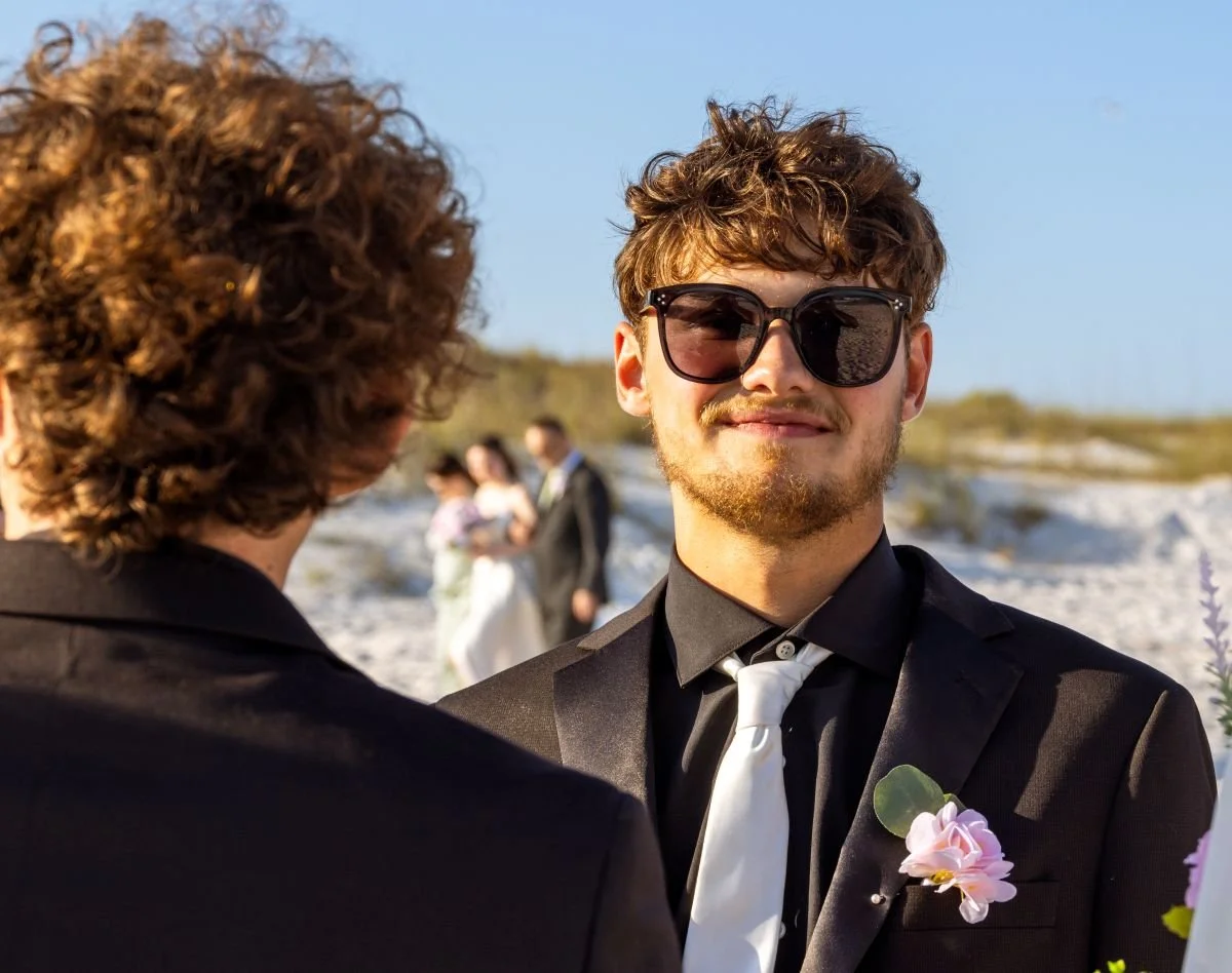 A young man with curly hair, sunglasses, and a black tuxedo with a white tie, smiling at an outdoor wedding ceremony on a beach, with other wedding guests in the background.