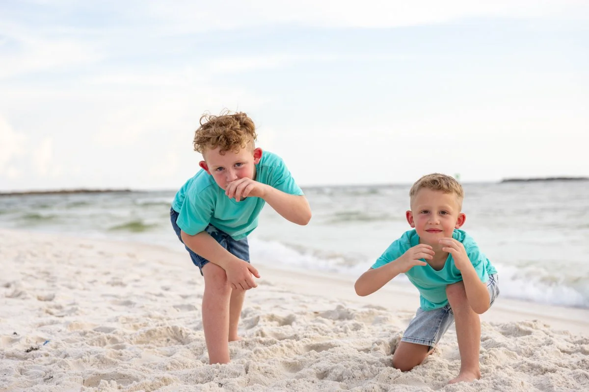 Two young boys playing in the sand on a beach, with ocean waves in the background and a cloudy sky overhead.