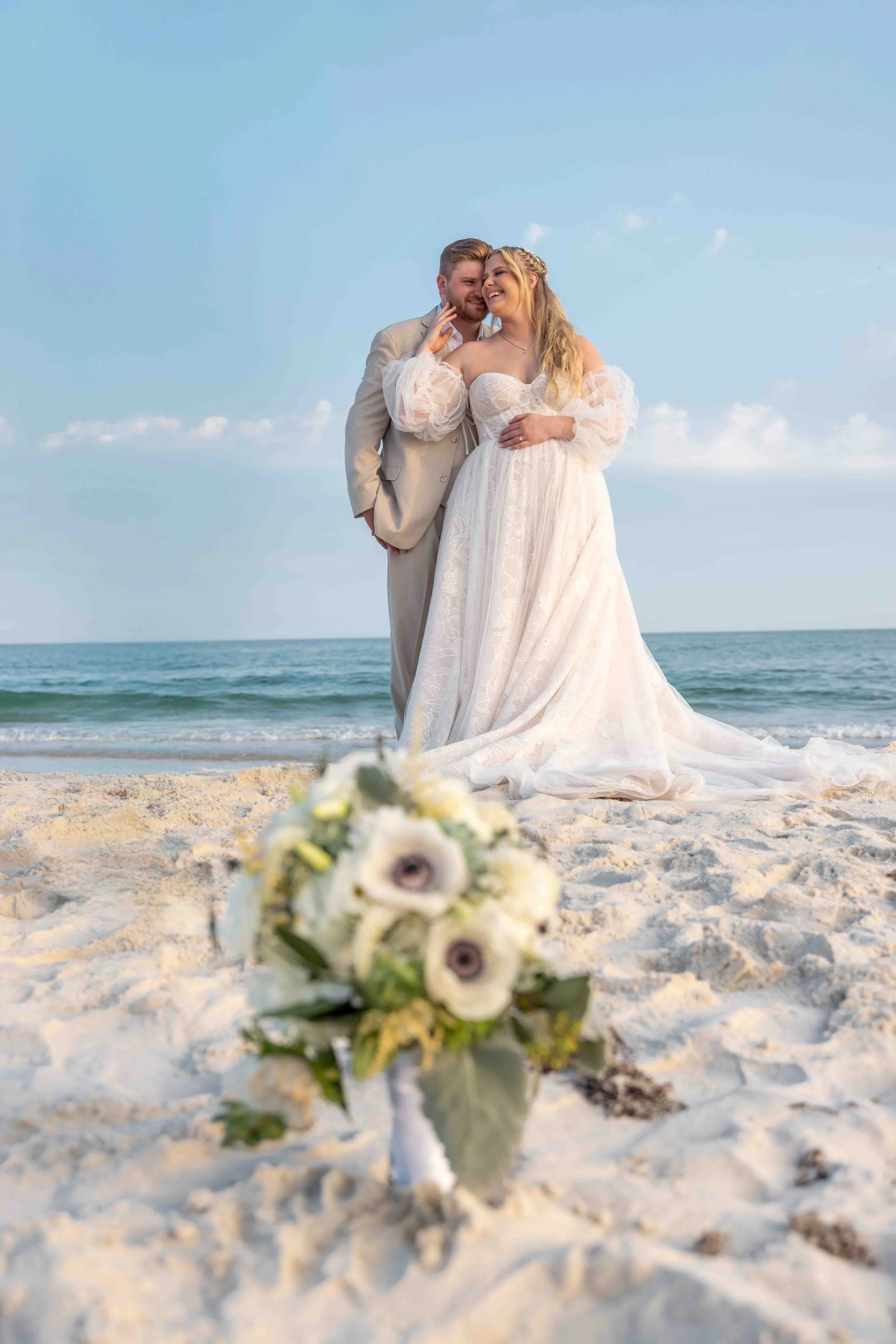 A bride and groom on a beach, smiling and leaning close to each other, with a bouquet of white and purple flowers in the foreground and the ocean and blue sky in the background.