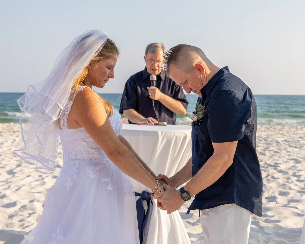 A beach wedding ceremony with a bride and groom holding hands, eyes closed, facing each other, while an officiant speaks into a microphone in the background.