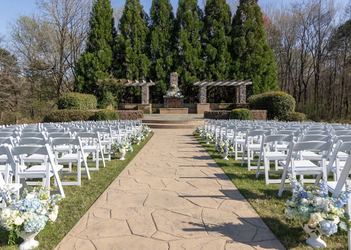 An outdoor wedding ceremony setup with white chairs arranged in rows on either side of a stone-paved aisle. The aisle is decorated with white and blue floral arrangements. At the front, a stage with a stone fireplace and decorative pillars is set aga