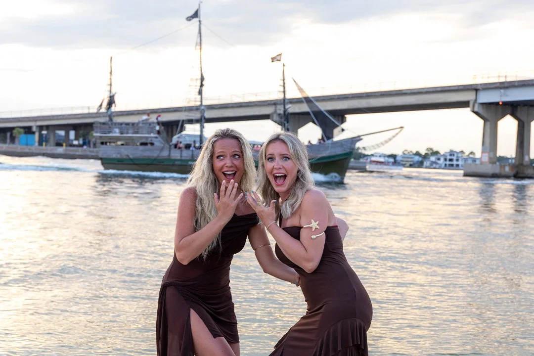 Two women in dark dresses standing on a waterfront with boats and a bridge in the background, smiling and posing playfully.