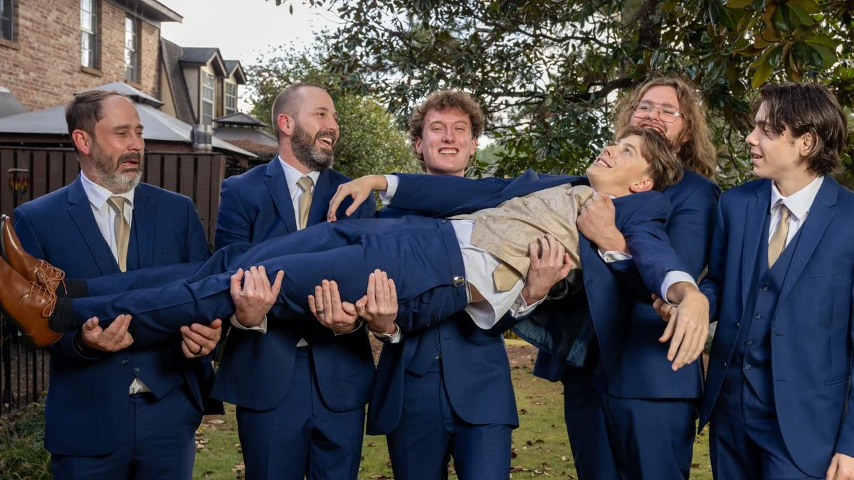 A group of men in suits holding a person horizontally in an outdoor setting with trees and houses in the background, during daytime.