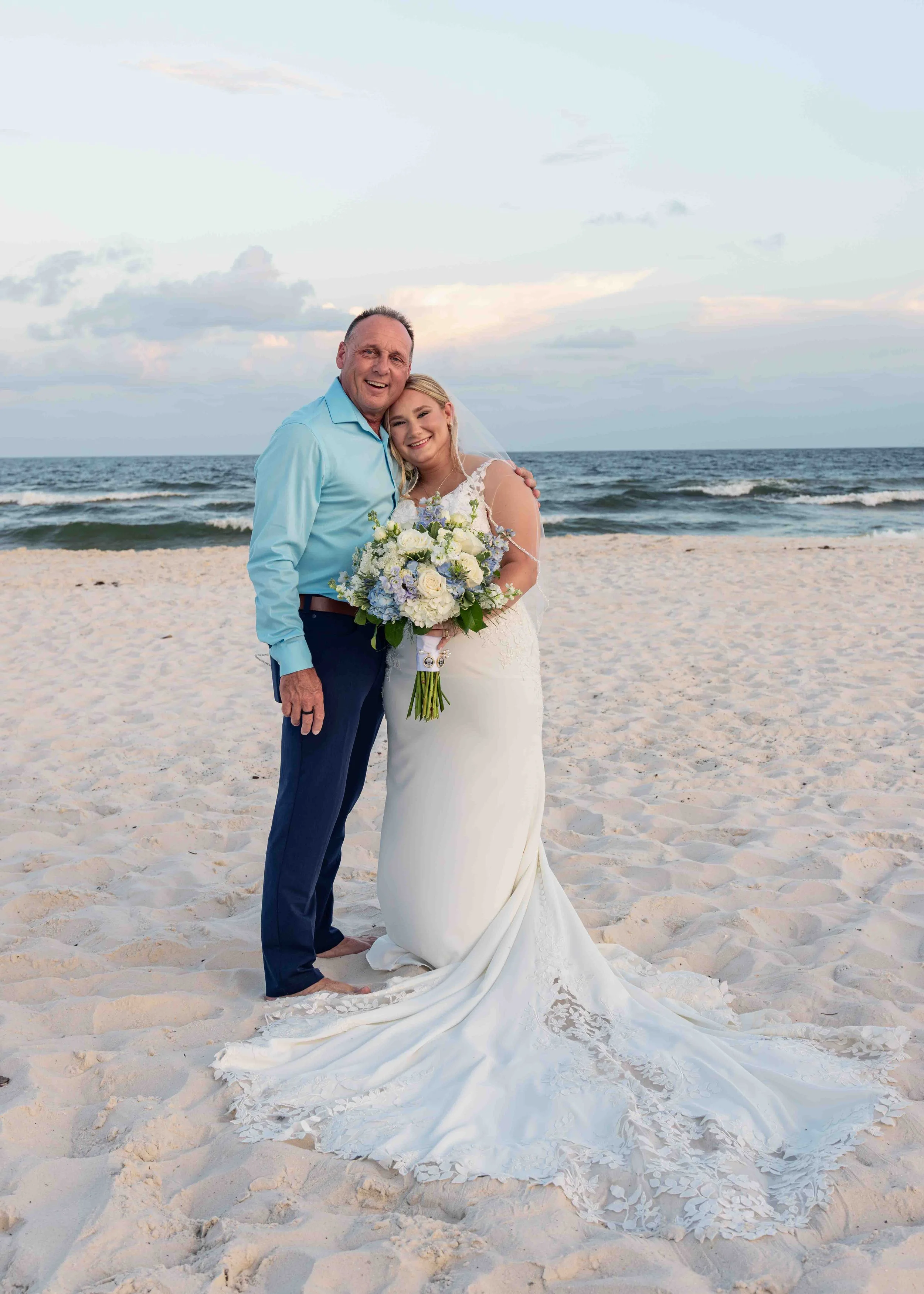 A man and a woman in wedding attire smiling and hugging on a sandy beach with the ocean and sky in the background.