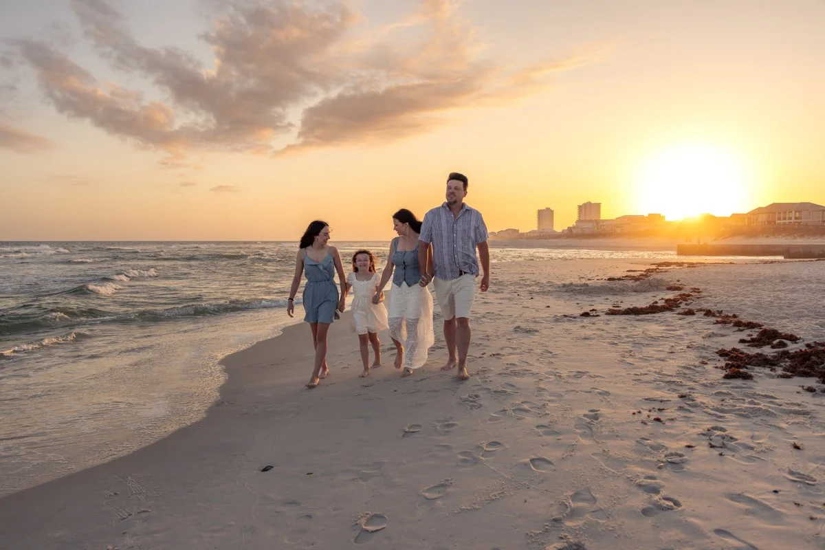 A family of four walking along the beach during sunset, with the ocean on the left and buildings in the background.