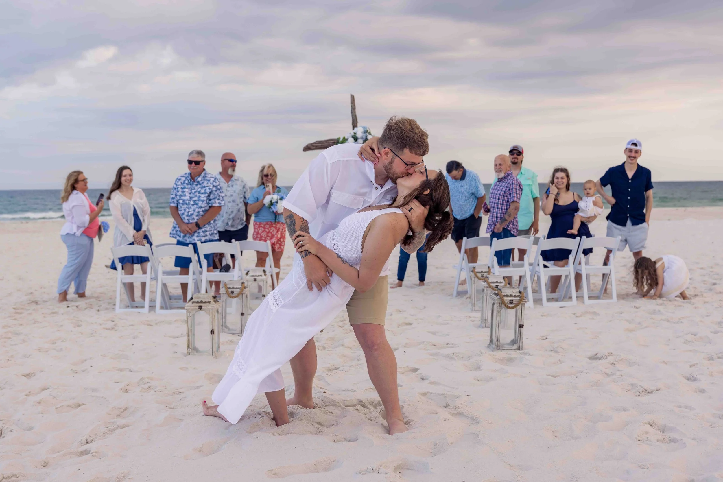 A wedding ceremony on the beach with a couple kissing, surrounded by friends and family. The groom is dipping the bride while kissing. The beach has chairs and lanterns, and the sky is cloudy.