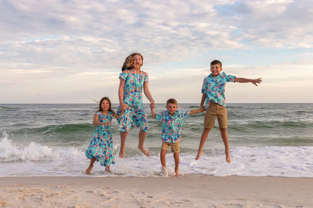 Group of five children wearing matching blue floral shirts and dresses, playing and jumping by the shoreline at the beach during sunset.