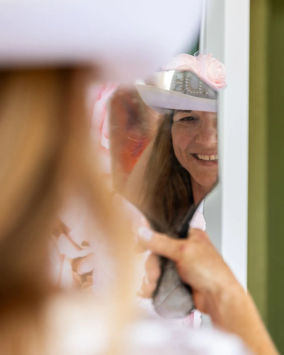 A woman smiles as she looks at herself in a mirror, wearing a tiara with flowers and the word 'BRIDE' on it.