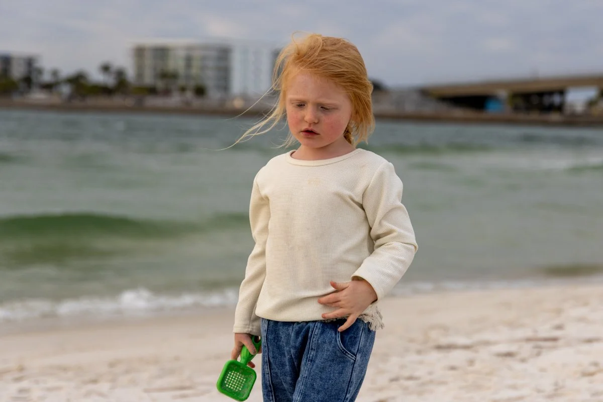 A young girl with red hair standing on a beach near the ocean, holding a green toy in her right hand, wearing a cream sweatshirt and blue jeans, with buildings and a bridge in the background.