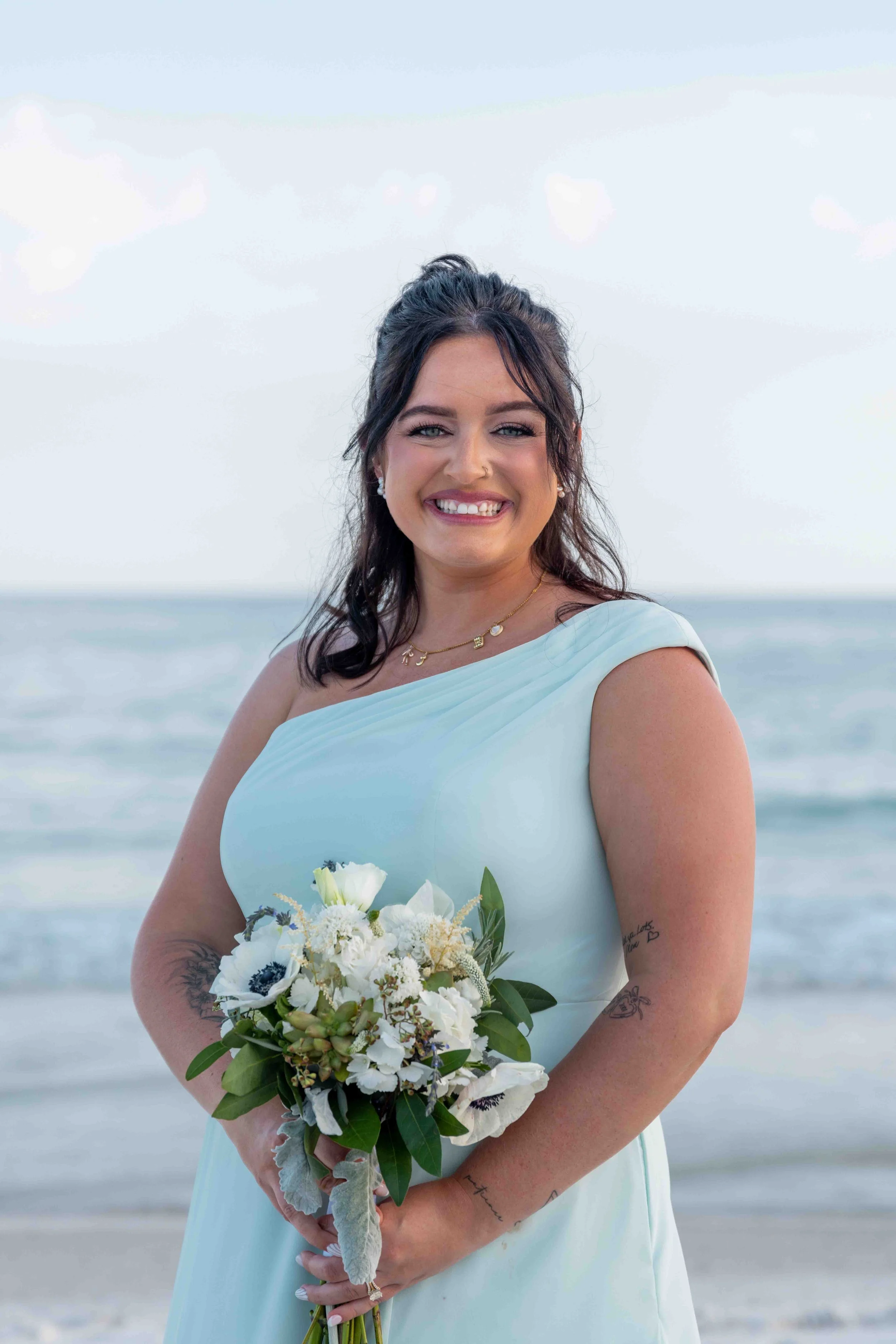 A smiling woman in a light blue dress holding a bouquet of white flowers on a beach with the ocean in the background.