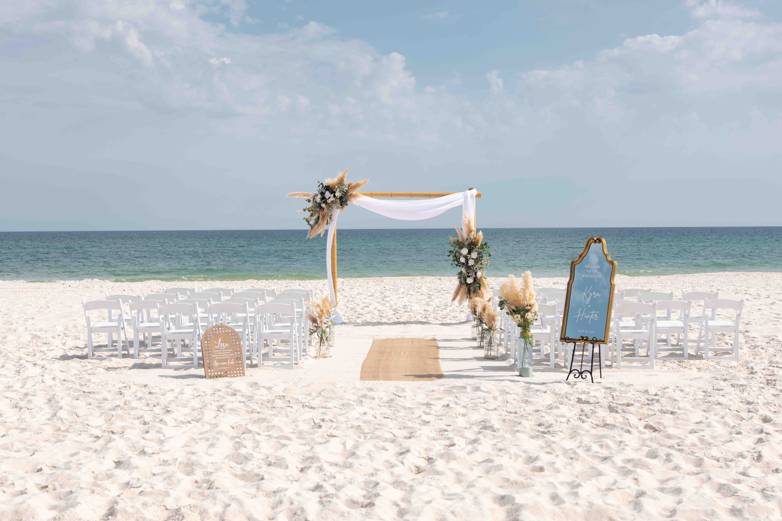 Beach wedding setup with white chairs arranged on sand facing the ocean, decorated arch with flowers and drapery, signs, and a clear sky.