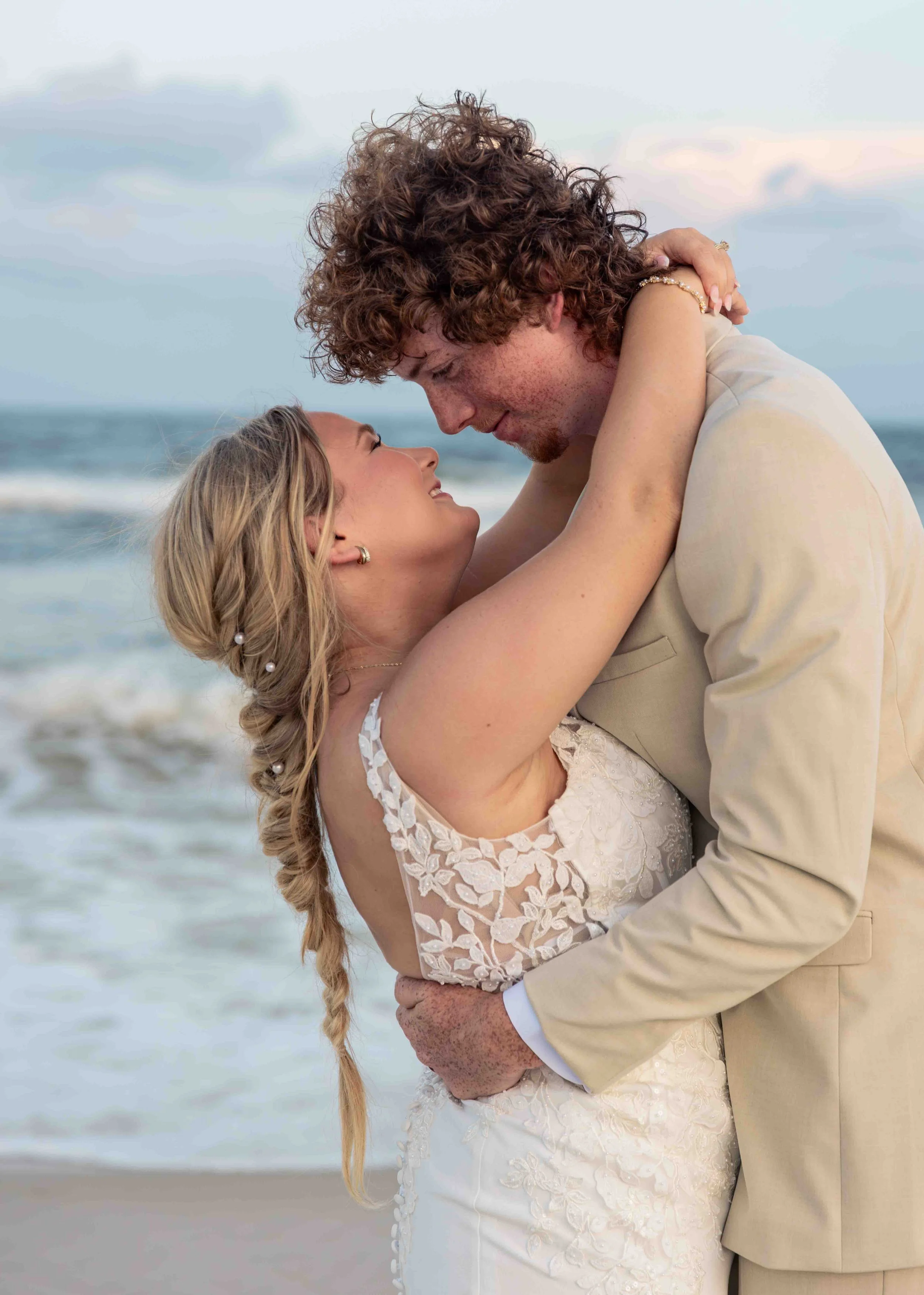 A bride and groom share an intimate moment on the beach, with the bride in a white lace wedding dress and the groom in a beige suit. The couple is holding each other close, smiling and gazing into each other's eyes. The ocean and sky are visible in t