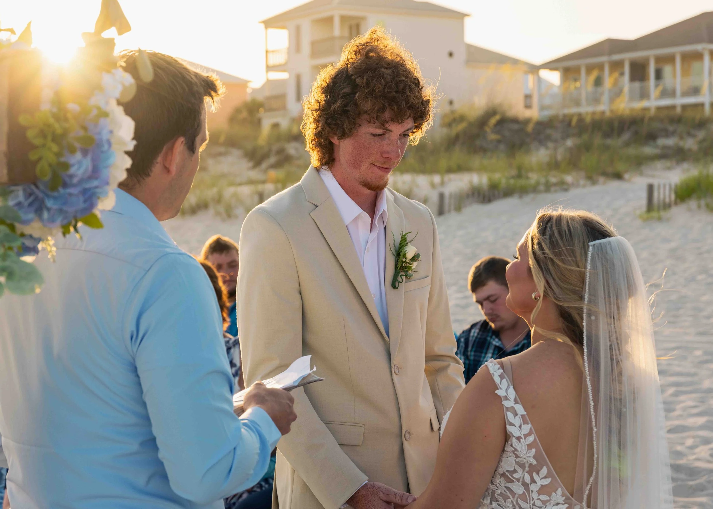 A wedding ceremony on the beach during sunset, with a groom and bride exchanging vows, surrounded by friends and family.