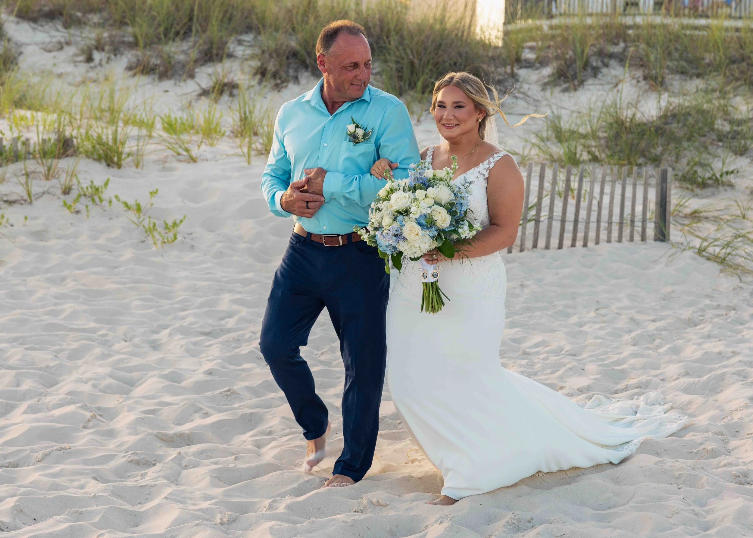 A bride holding a bouquet of white and blue flowers walking on the beach with an older man, possibly her father, during a wedding ceremony. The man is dressed in a light blue shirt and dark pants, the bride wears a white lace wedding dress with a flo