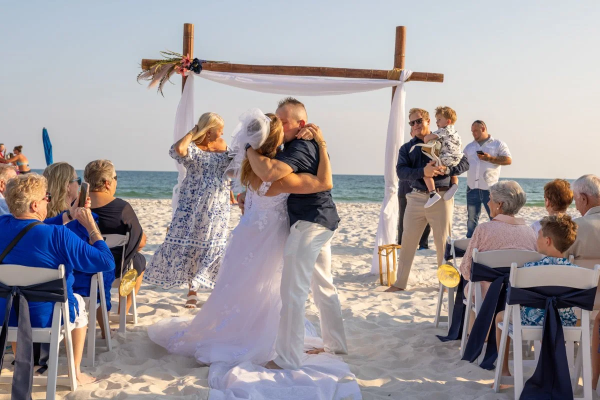 A beach wedding ceremony with a bride and groom kissing under a wooden arch, surrounded by seated guests and spectators, with the ocean in the background.