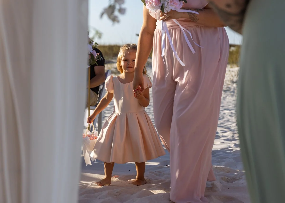 A young girl in a peach-colored dress holding a small basket, seen during a wedding on the beach. She is looking through draped fabric at adults, and there are flowers and other people in the background.