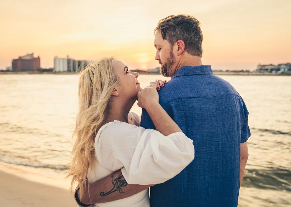 A couple embraces on a beach at sunset, with city buildings in the background.