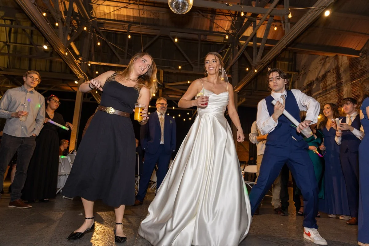 People dancing and celebrating at a wedding reception in an industrial-style venue with string lights and a disco ball.