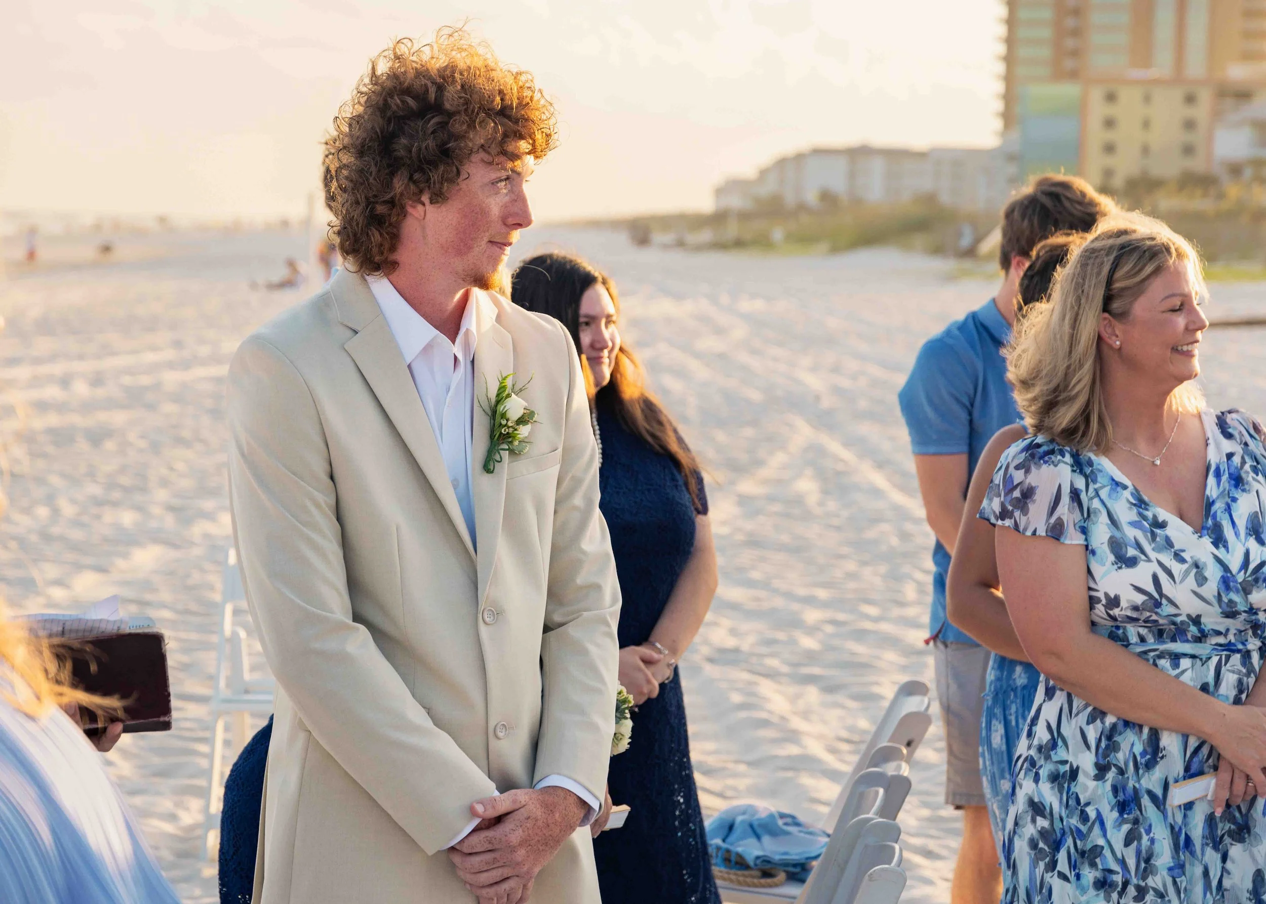 A man in a beige suit with a white shirt, standing with his hands clasped, during an outdoor wedding ceremony on the beach at sunset. He has curly hair and a boutonniere pinned to his lapel, with other guests visible behind him.