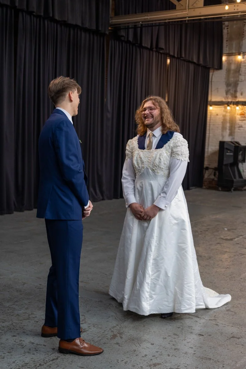 A wedding ceremony with a man in a dark blue suit and a woman in a white gown with lace details, standing and smiling at each other inside a venue with black curtains and exposed brick walls.