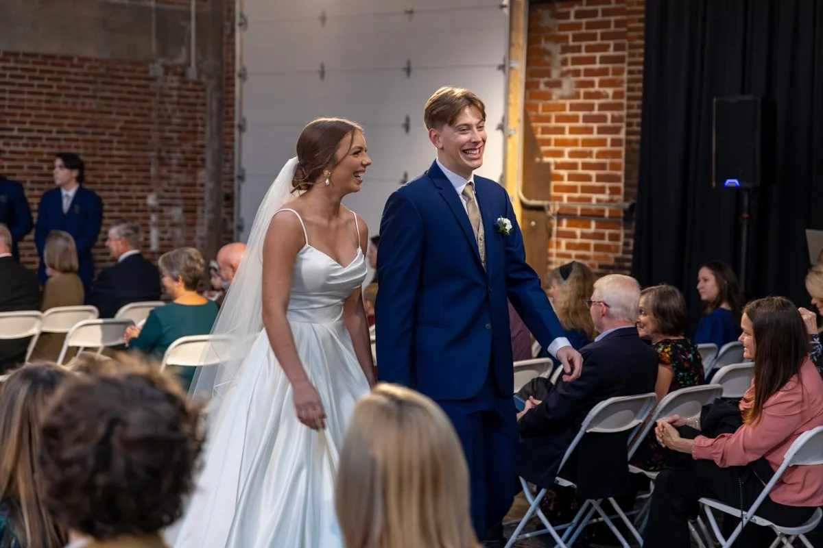 Bride and groom smiling as they walk down the aisle at their wedding ceremony, surrounded by seated guests.