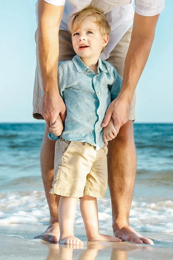 A young boy holding his father's hands on a beach with ocean waves in the background.