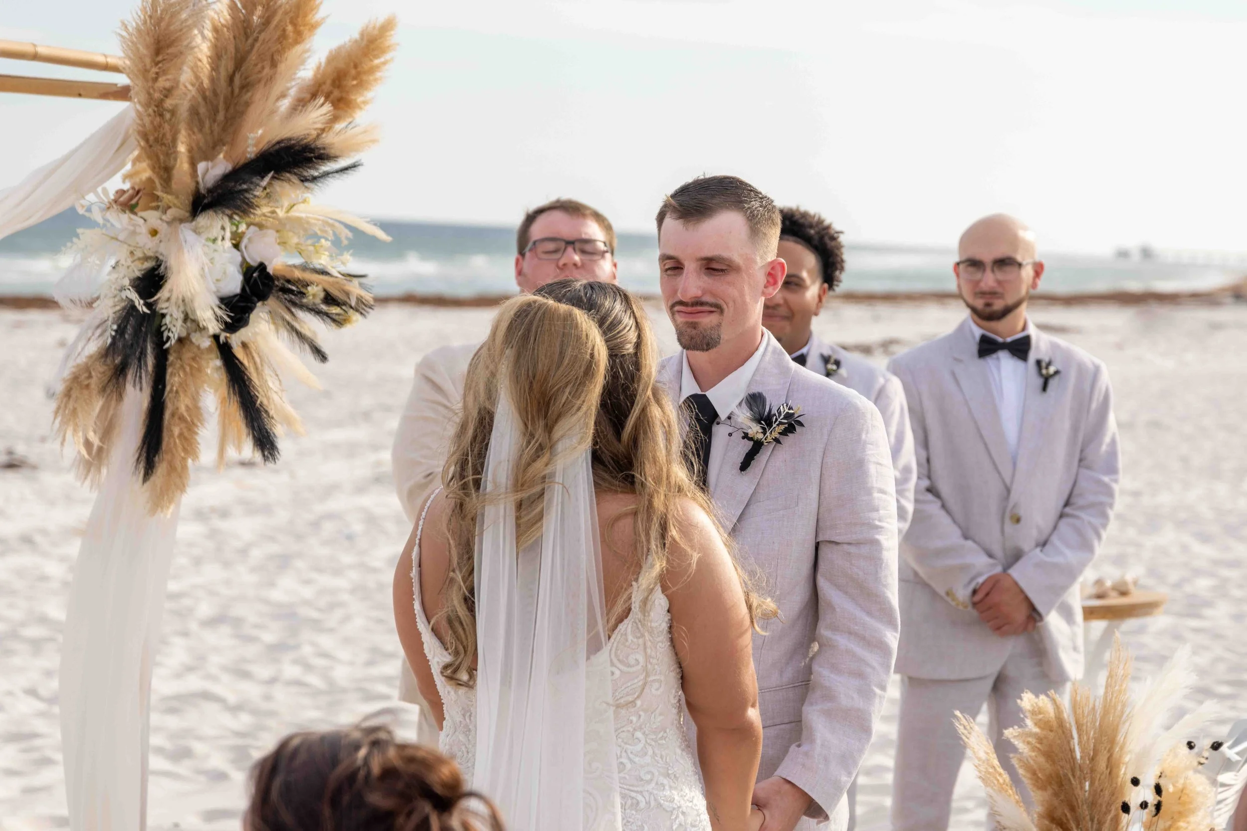 A beach wedding ceremony with a groom and bride facing each other, surrounded by groomsmen in light suits and a decorated altar with pampas grass, on a sandy beach with the ocean in the background.