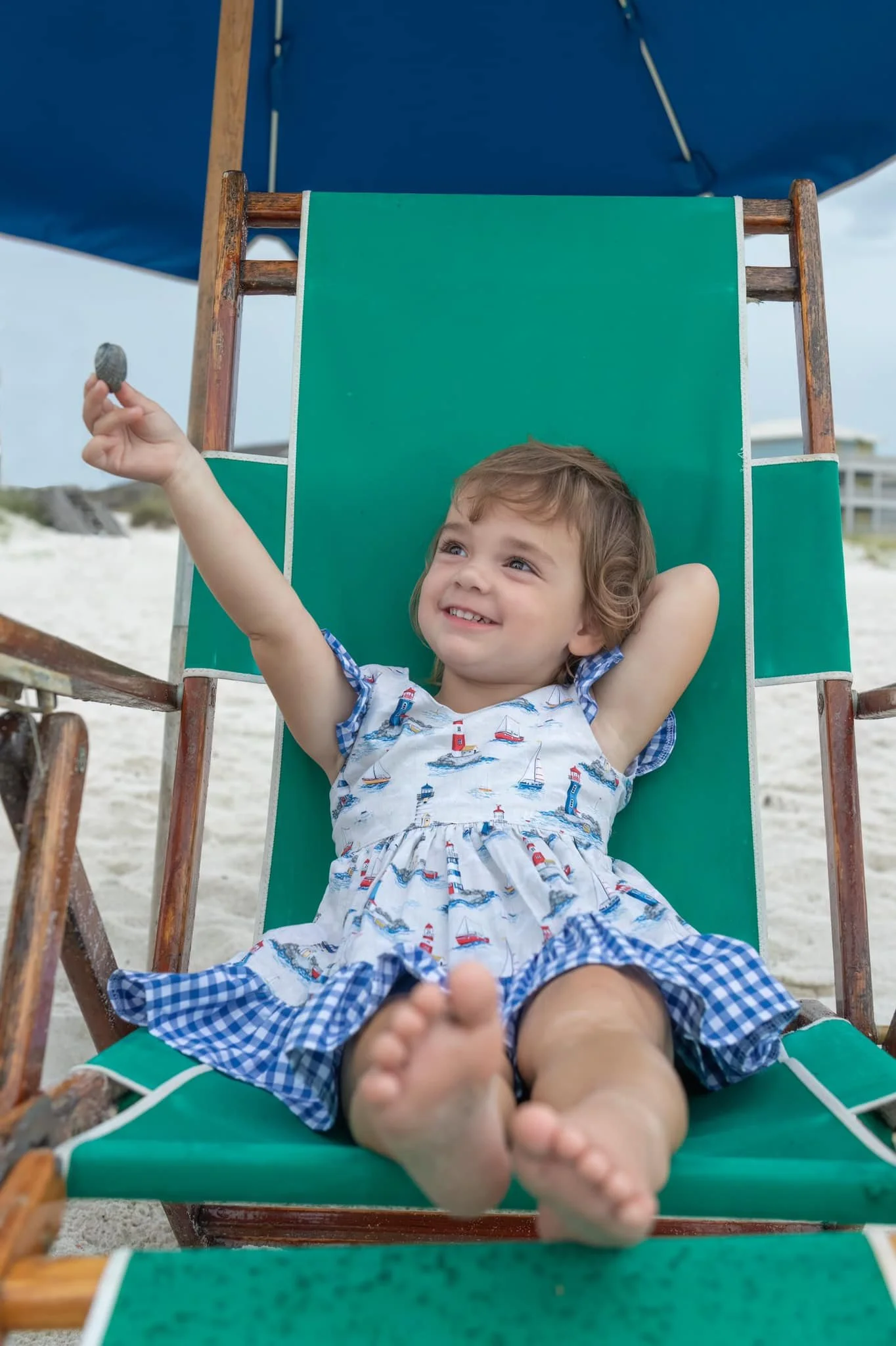 A young boy with short, curly hair lying on a green beach lounge chair at the beach, smiling and holding a small shell in his hand, with a large beach umbrella above him.
