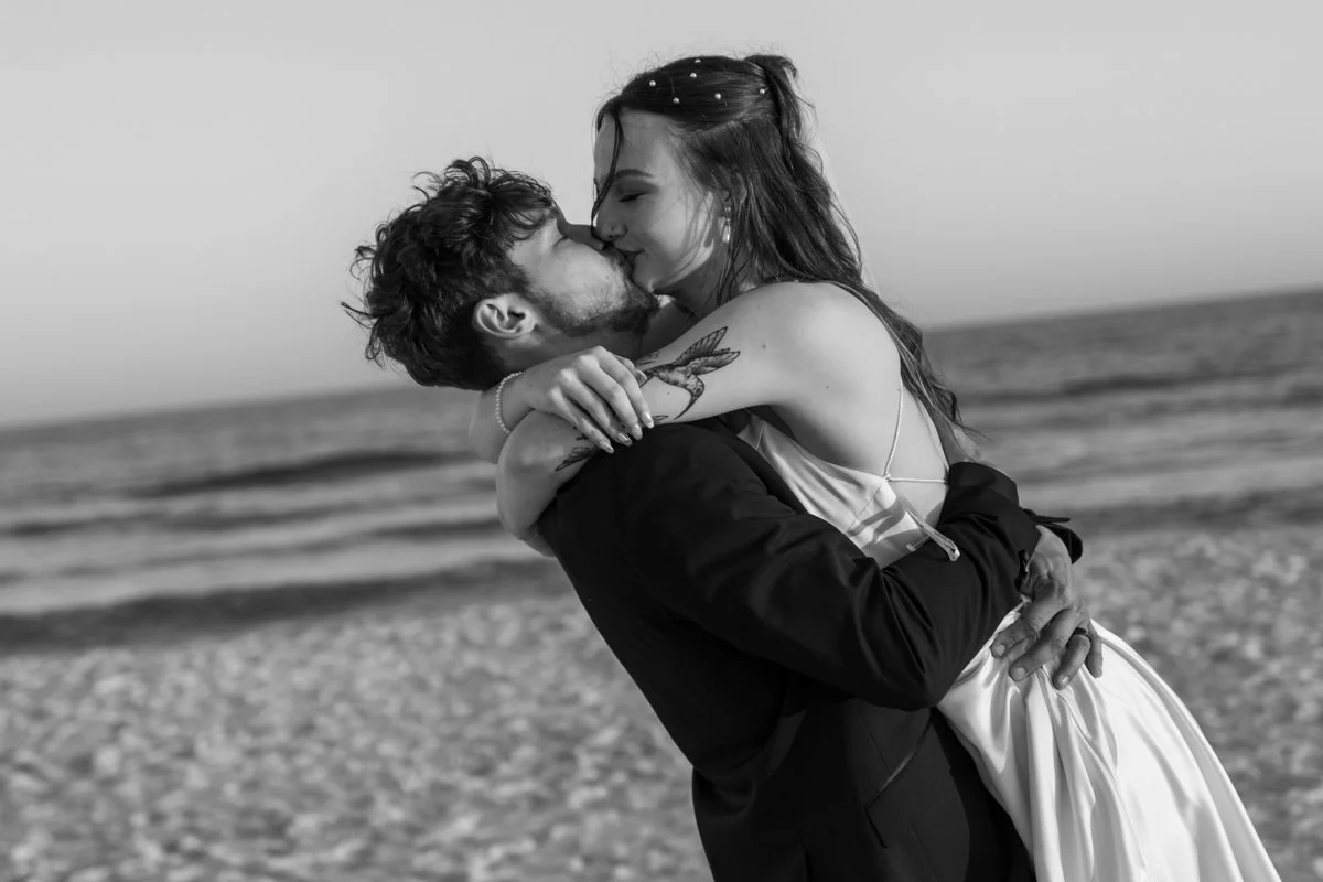 A couple kissing on the beach, the man lifting the woman in his arms, black and white photo, ocean in the background.