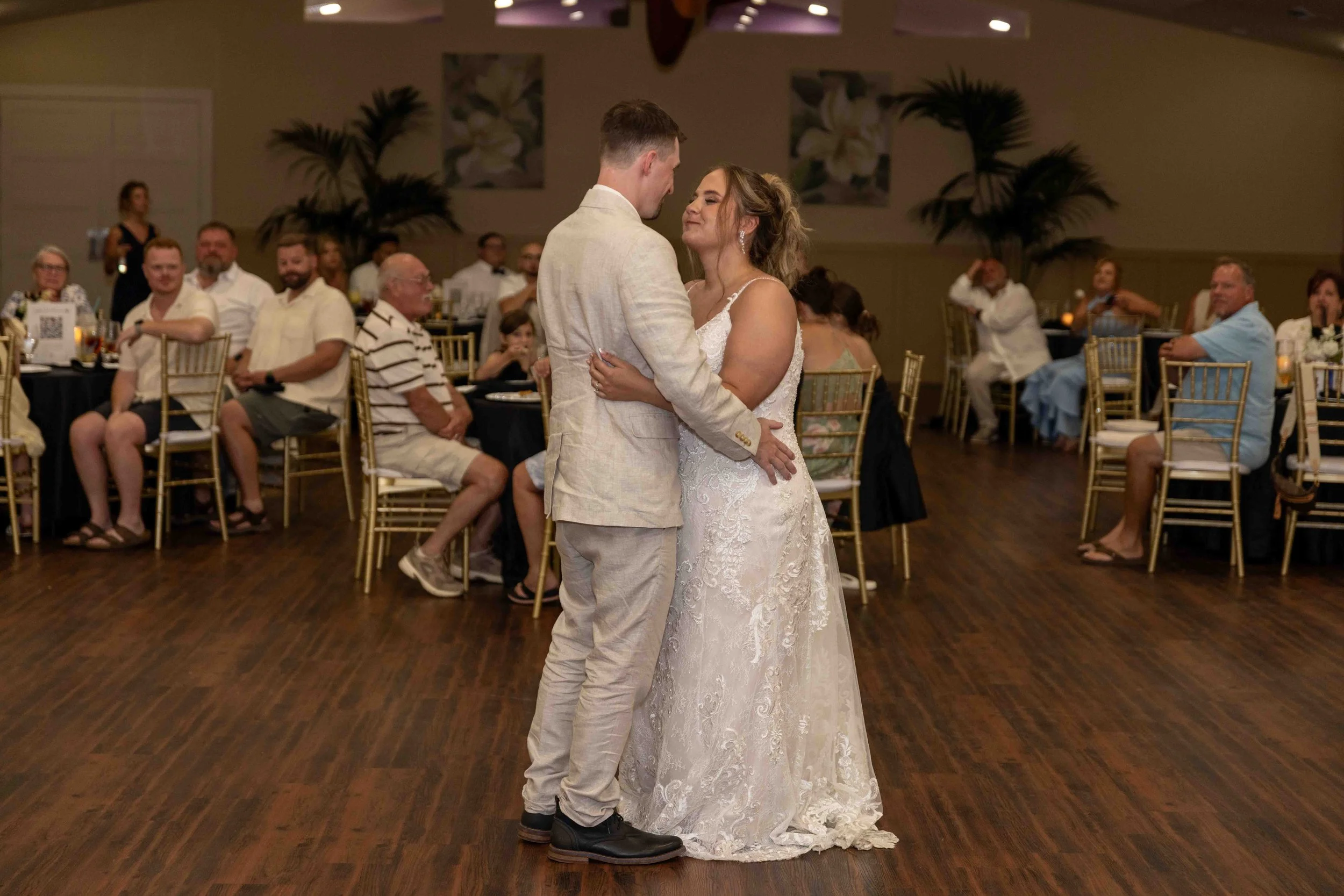 A bride and groom are dancing in the center of a reception hall, surrounded by seated guests watching and smiling.