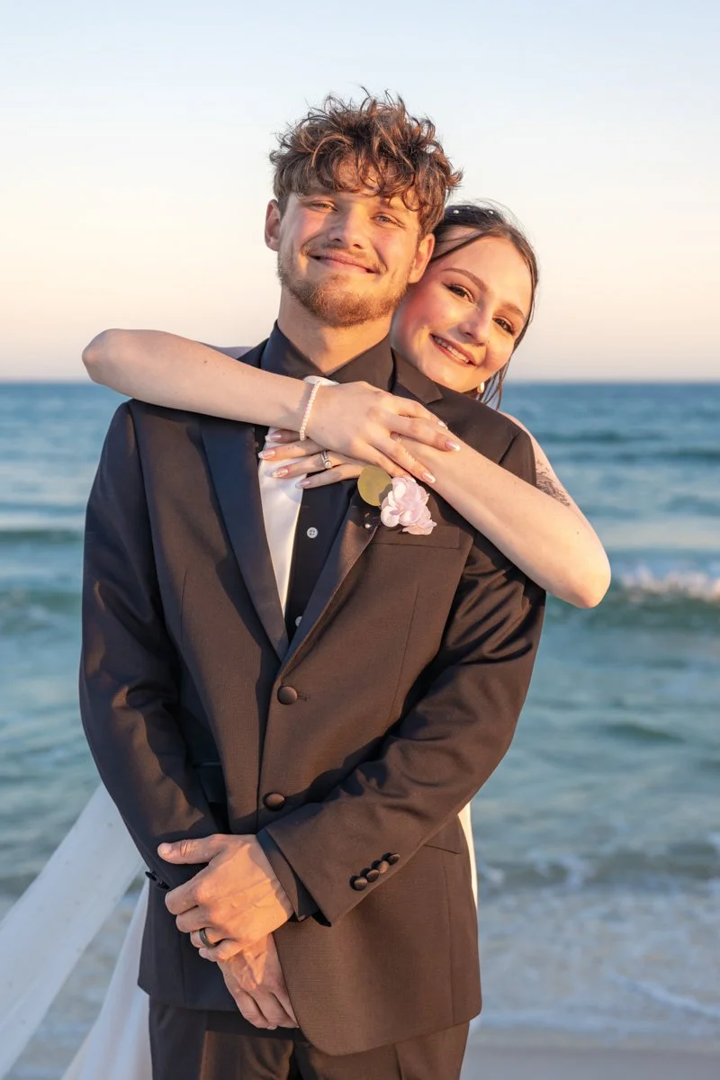 A newlywed couple on the beach, smiling and embracing during sunset, with the ocean in the background.