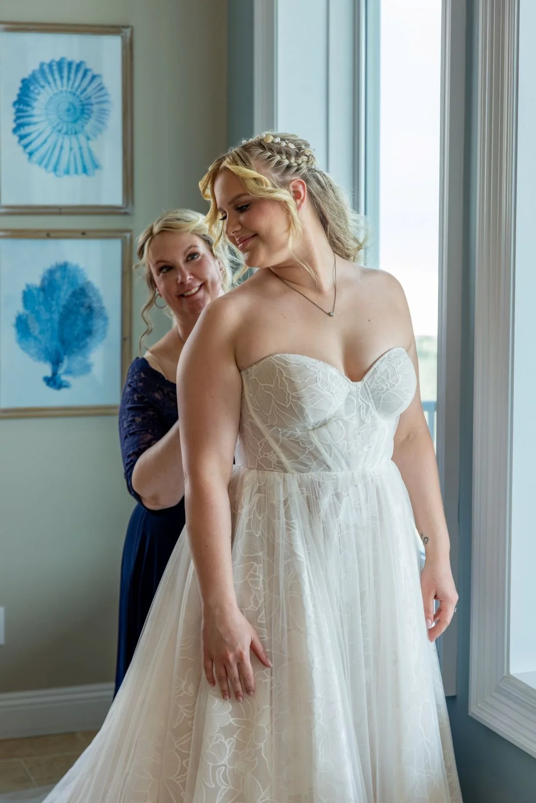 A bride in a white lace strapless wedding dress getting ready with the help of a woman, possibly her mother or a friend, in a room with framed blue shell artwork on the walls and a large window.