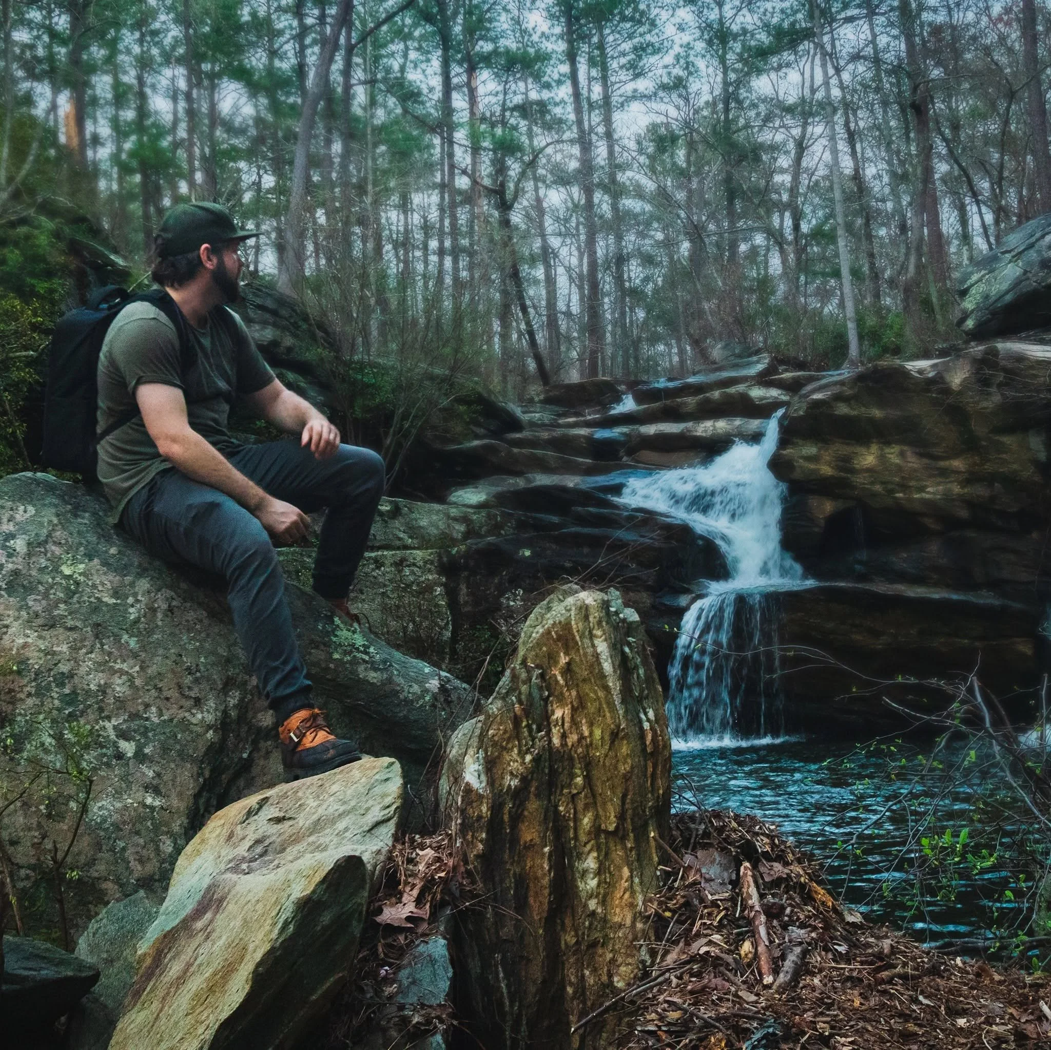 A man with a beard, wearing a cap and backpack, sitting on rocks by a stream in a dense forest with tall trees and moss-covered rocks.
