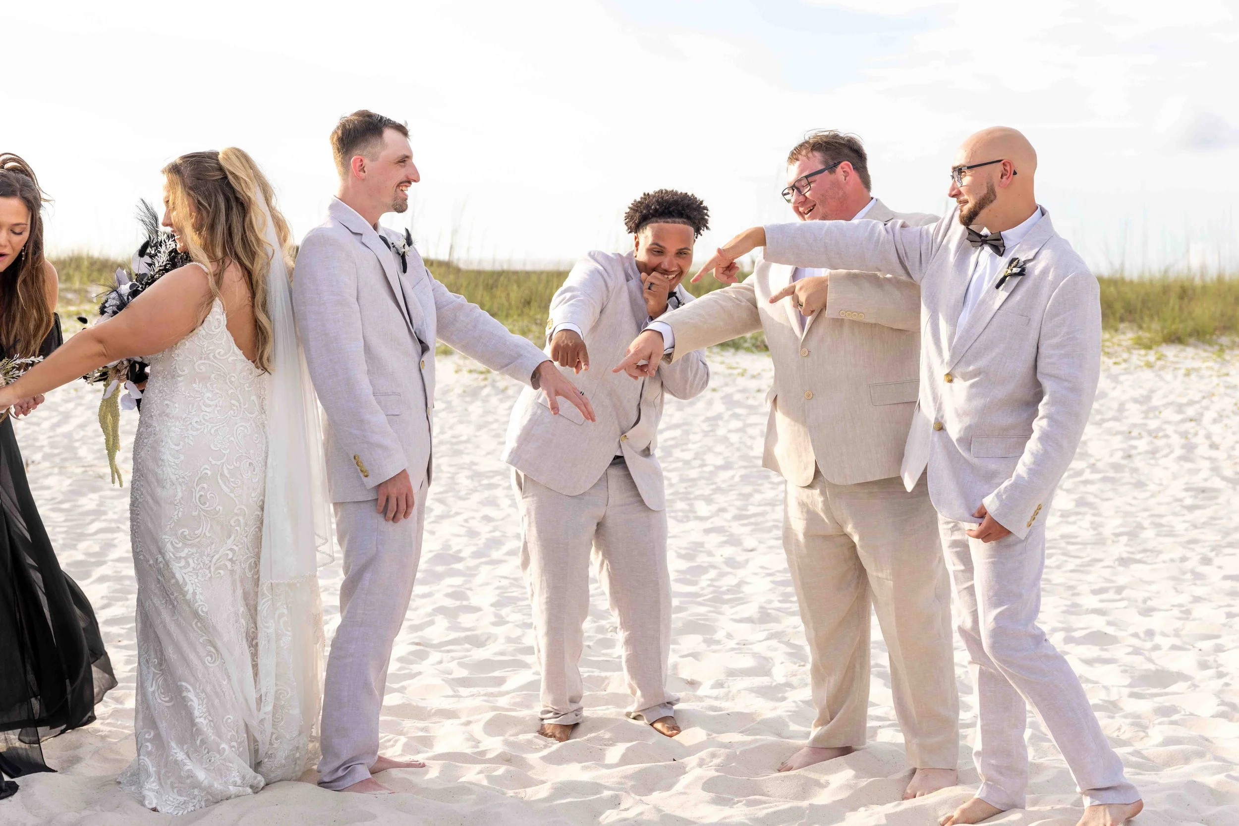 A group of wedding guests, dressed in light-colored and beige clothing, standing on a sandy beach, smiling and making a toast.
