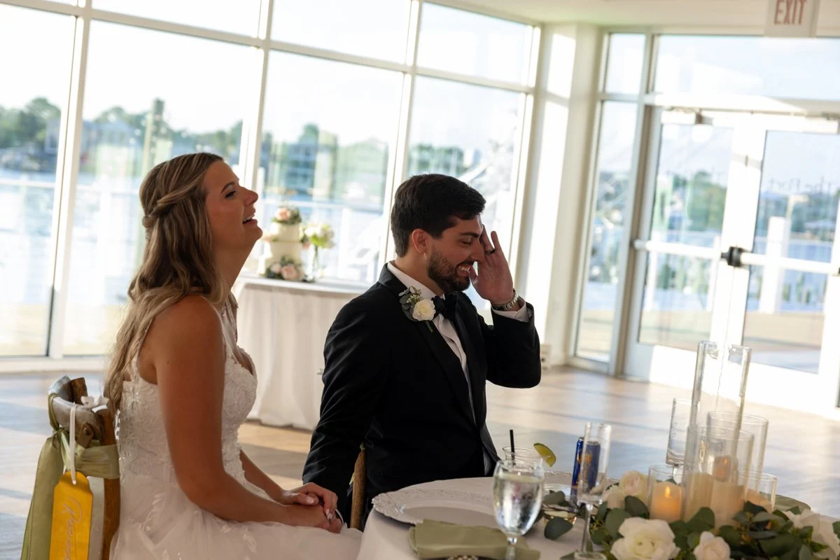 A bride and groom sitting at a table during a wedding reception, with the bride smiling and the groom appearing emotional, inside a glass-walled venue with waterfront views.
