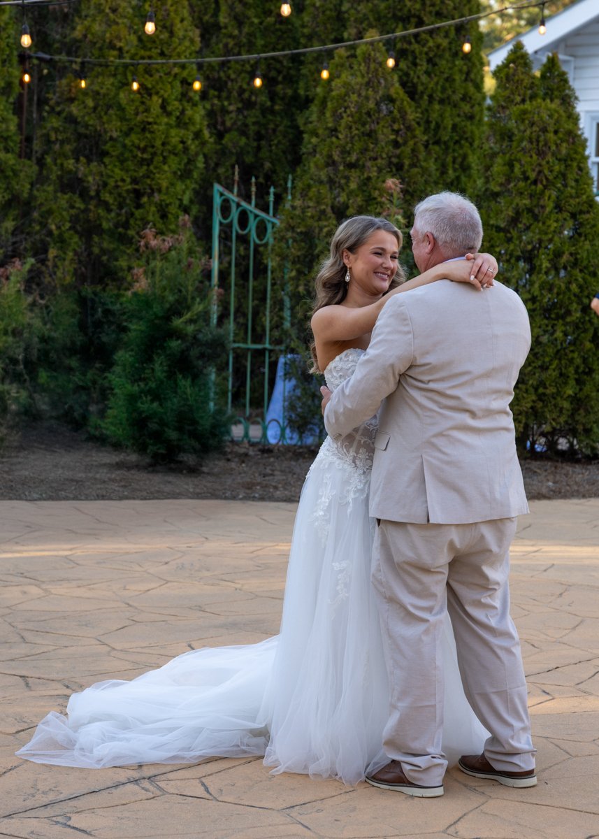A bride in a white wedding dress dancing with an older man in a light-colored suit outdoors. String lights hang overhead, and there are green trees and bushes in the background.