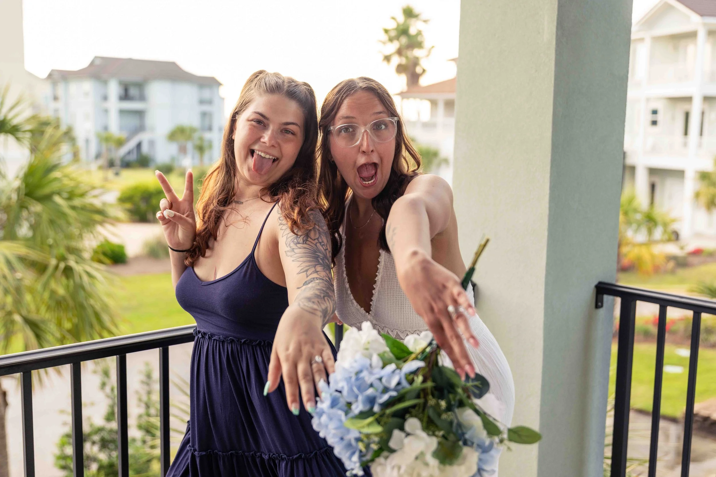 Two women celebrating a special occasion on a balcony with flowers, one wearing glasses, and both smiling and reaching toward the camera.
