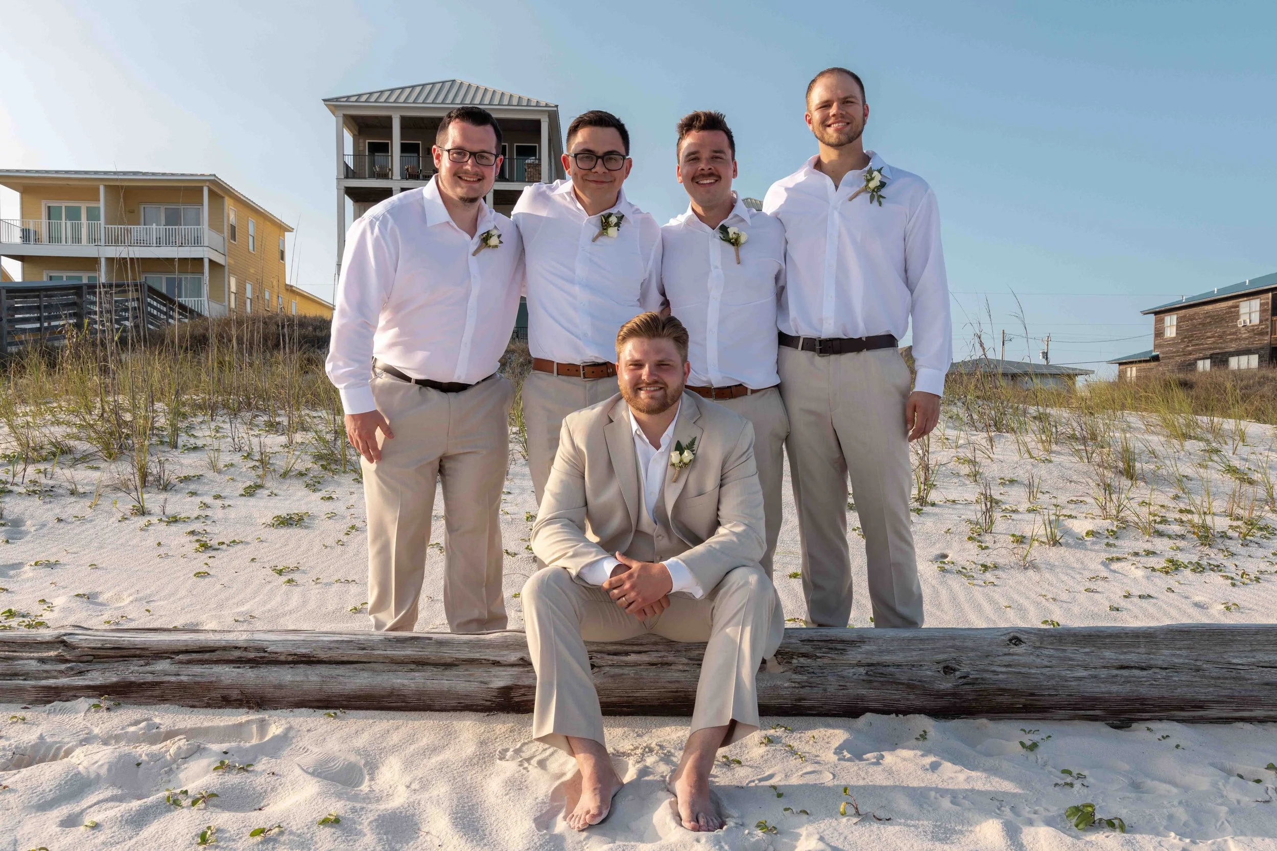 Group of five men in white shirts and beige pants posing on a sandy beach with houses in the background.