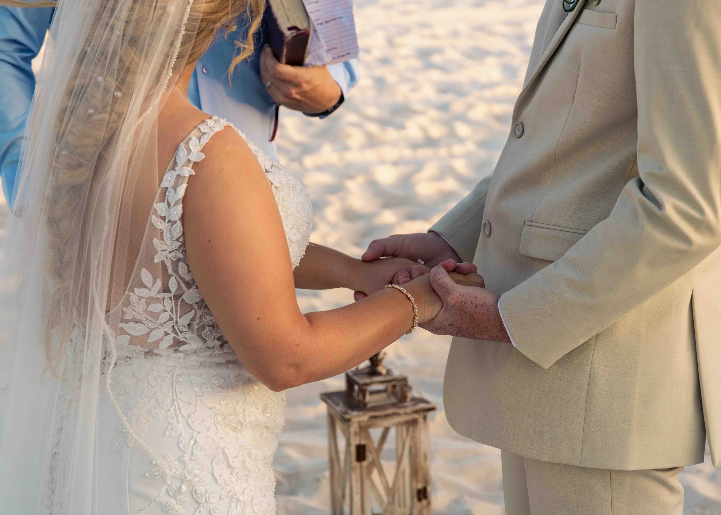 Close-up of a couple exchanging wedding vows outdoors on a sandy beach, with a officiant in the background holding a book, during sunset or late afternoon.