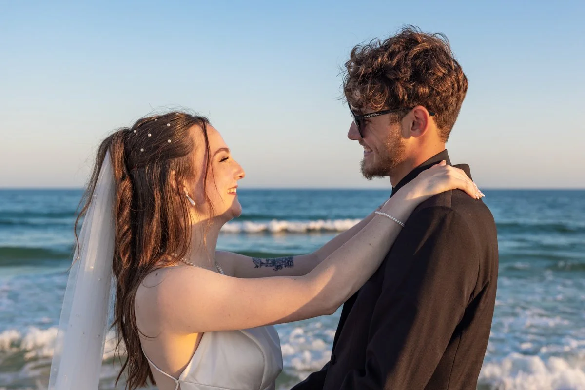 A newlywed couple stands on the beach embracing each other, with the ocean waves in the background, on a sunny day.