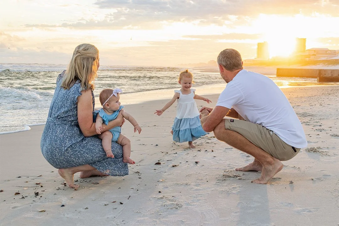 A family of four on the beach during sunset, with two young girls, one of whom is being held by a woman, and the other girl is playing with a man. The family appears to be enjoying a joyful moment by the ocean.