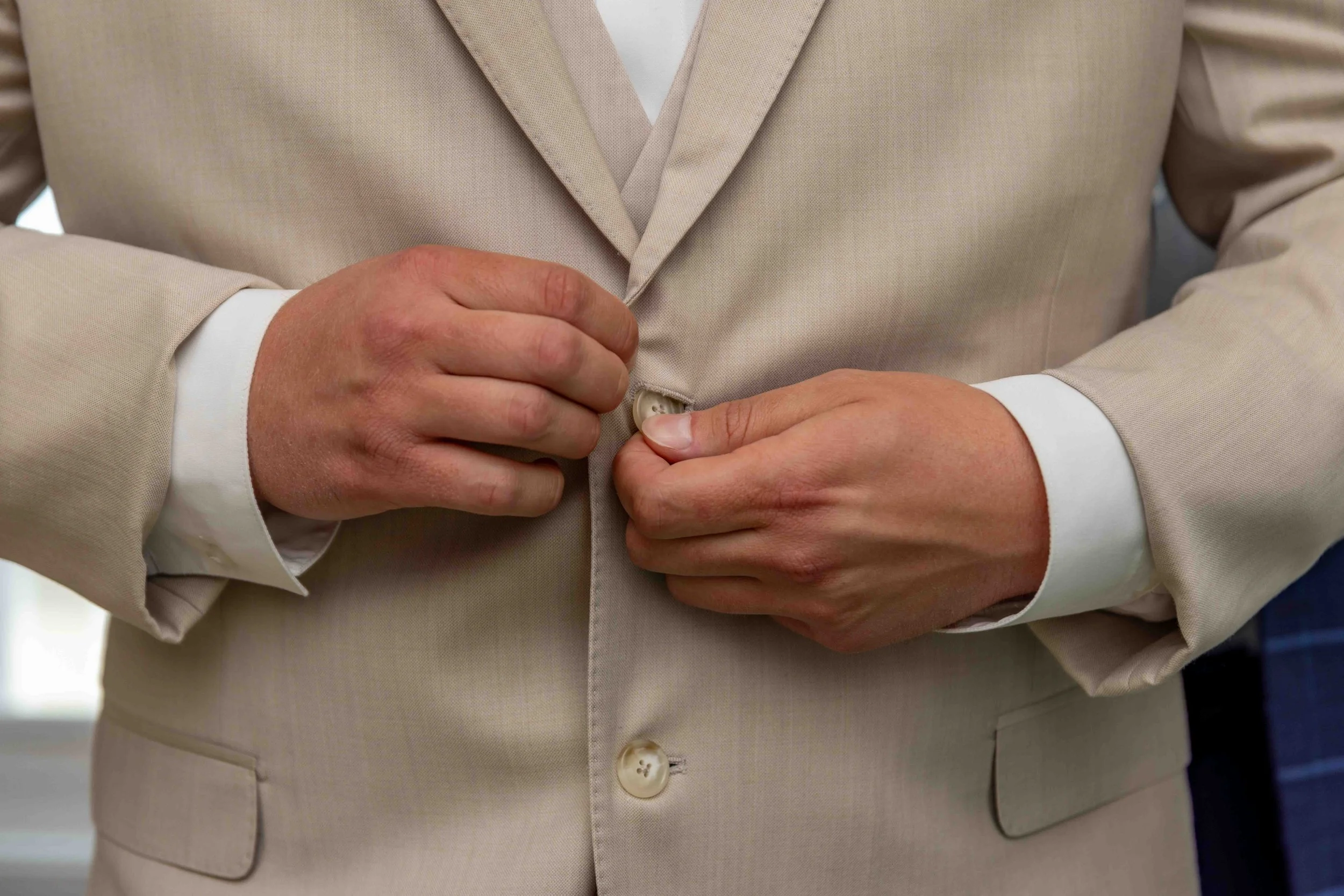 Person wearing a beige suit and white shirt buttoning the suit jacket.