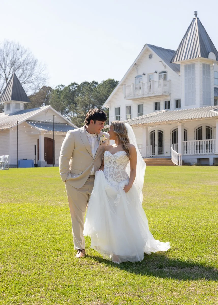 Bride and groom in wedding attire standing on a lush green lawn in front of a white mansion with Victorian architectural style.