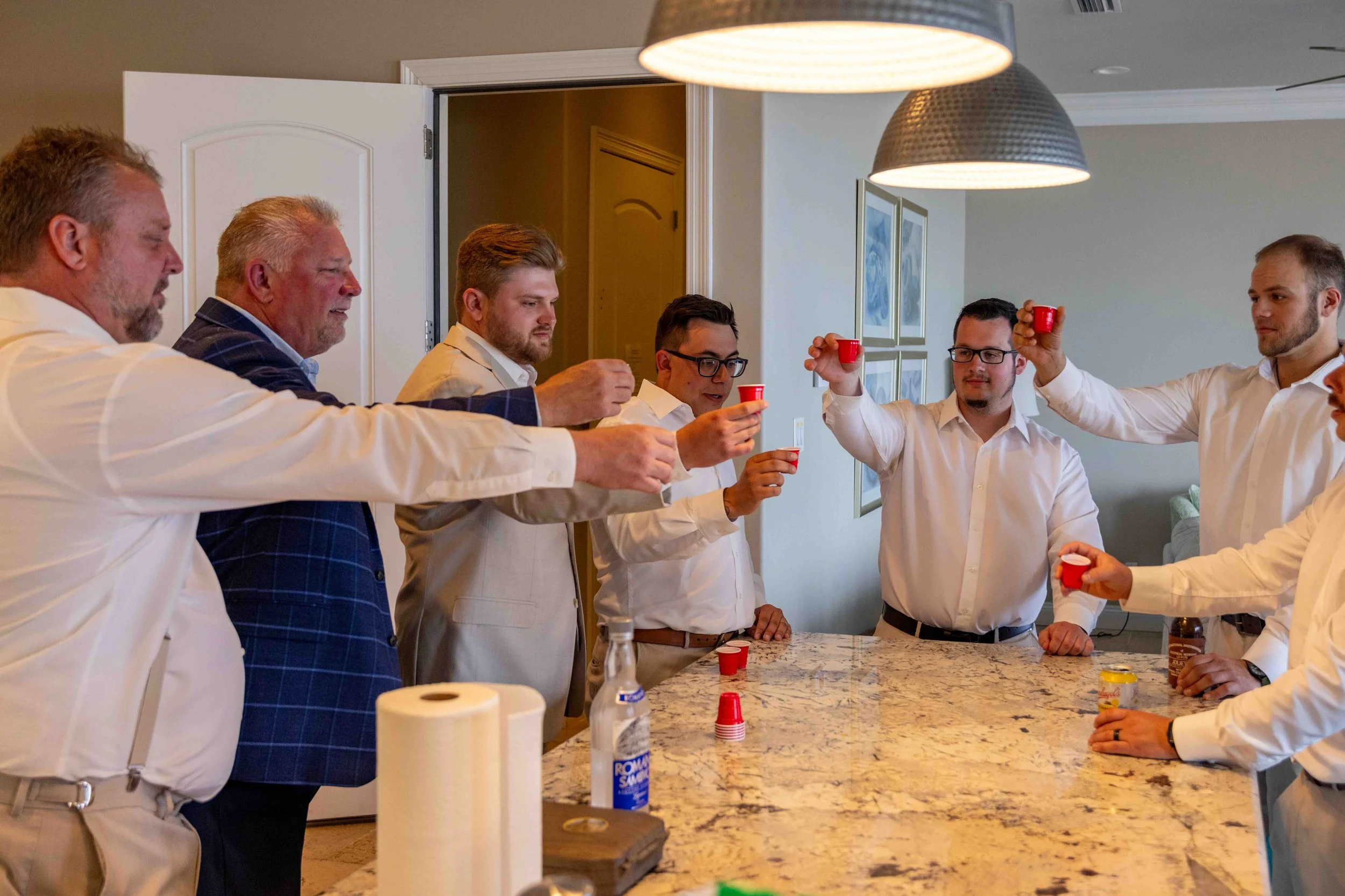 A group of seven men in business attire raising red shot glasses in a toast in a modern kitchen with a granite island and contemporary lighting fixtures.
