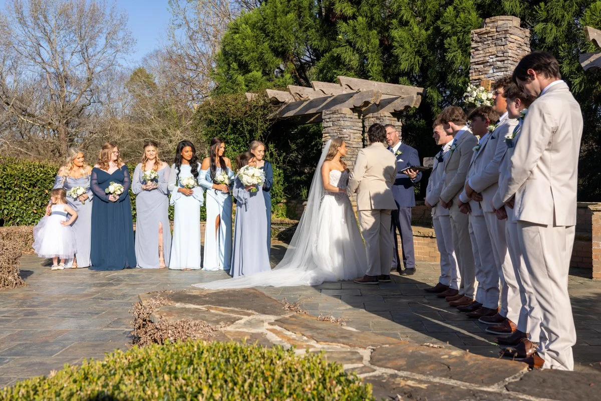 A wedding ceremony outside with the bride and groom standing in front of an officiant, surrounded by bridesmaids and groomsmen, under a wooden and stone archway with lush greenery and trees in the background.