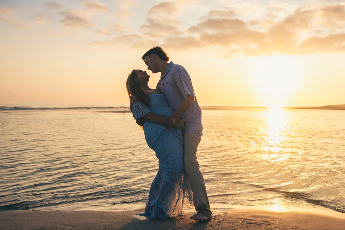 A couple stands on the beach at sunset, embracing and looking into each other's eyes.