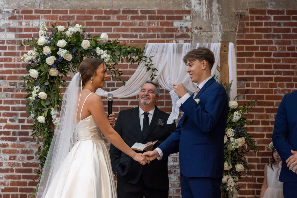 Bride and groom holding hands during their wedding ceremony, exchanging vows, with officiant smiling in front of a floral arch against a brick wall.