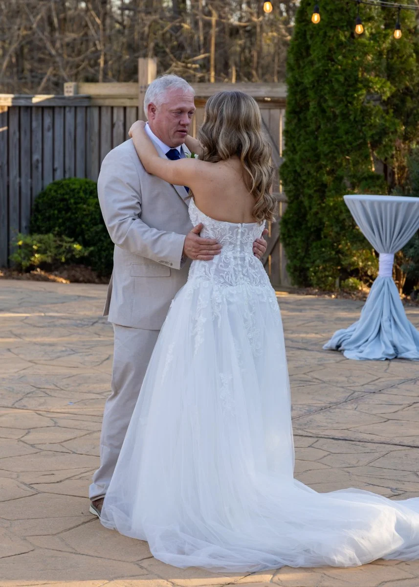 A bride and groom are dancing outdoors at a wedding reception. The bride is wearing a white wedding dress with lace details, and the groom is in a light-colored suit. They are holding each other closely, sharing a tender moment.