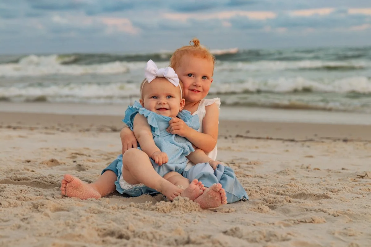 Two young children, a girl and a baby, sitting on the sandy beach with ocean waves in the background. The girl has curly red hair and wears a white dress, while the baby has blue eyes, wears a blue dress, and has a large white bow in her hair. They a