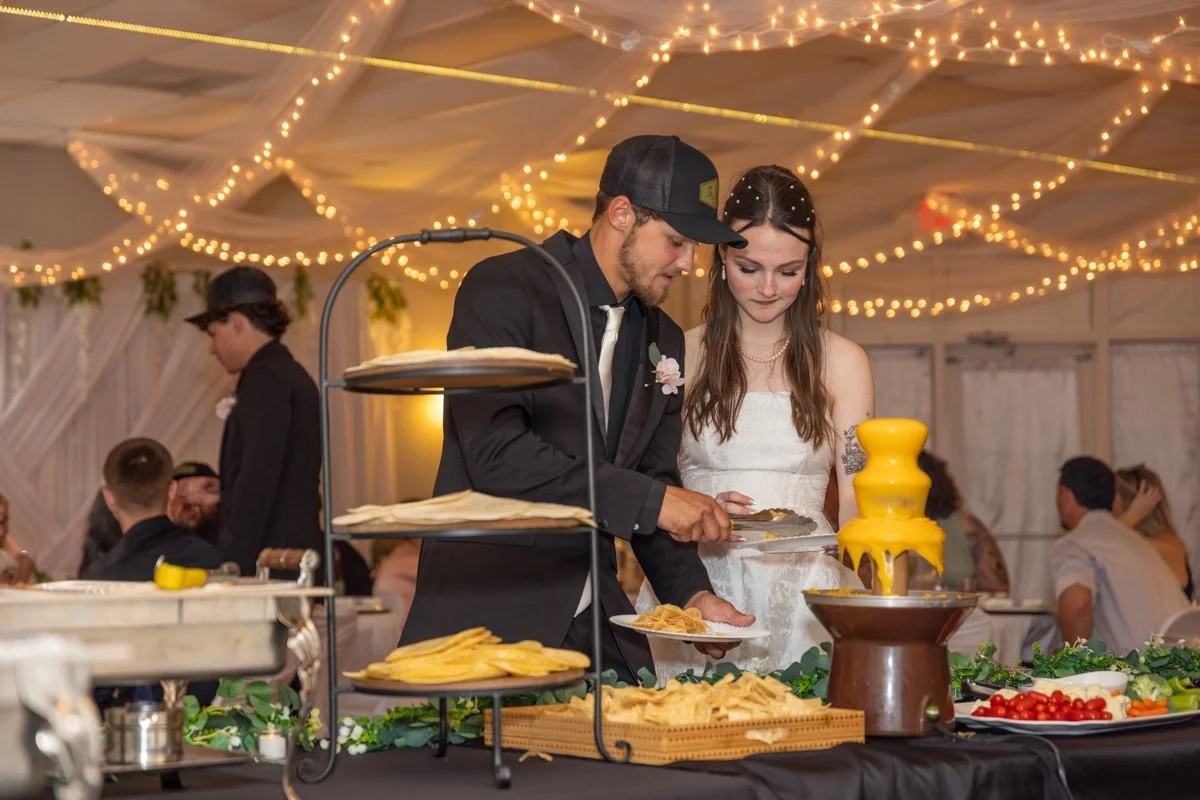 A bride and groom serve themselves at a wedding reception buffet with string lights on the ceiling, with other guests visible in the background.